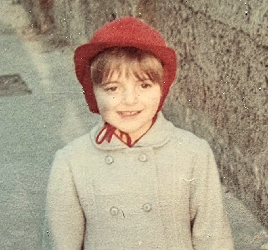 Lachendes Mädchen mit rotem Hut und hellem Mantel in einer Dorfstraße.