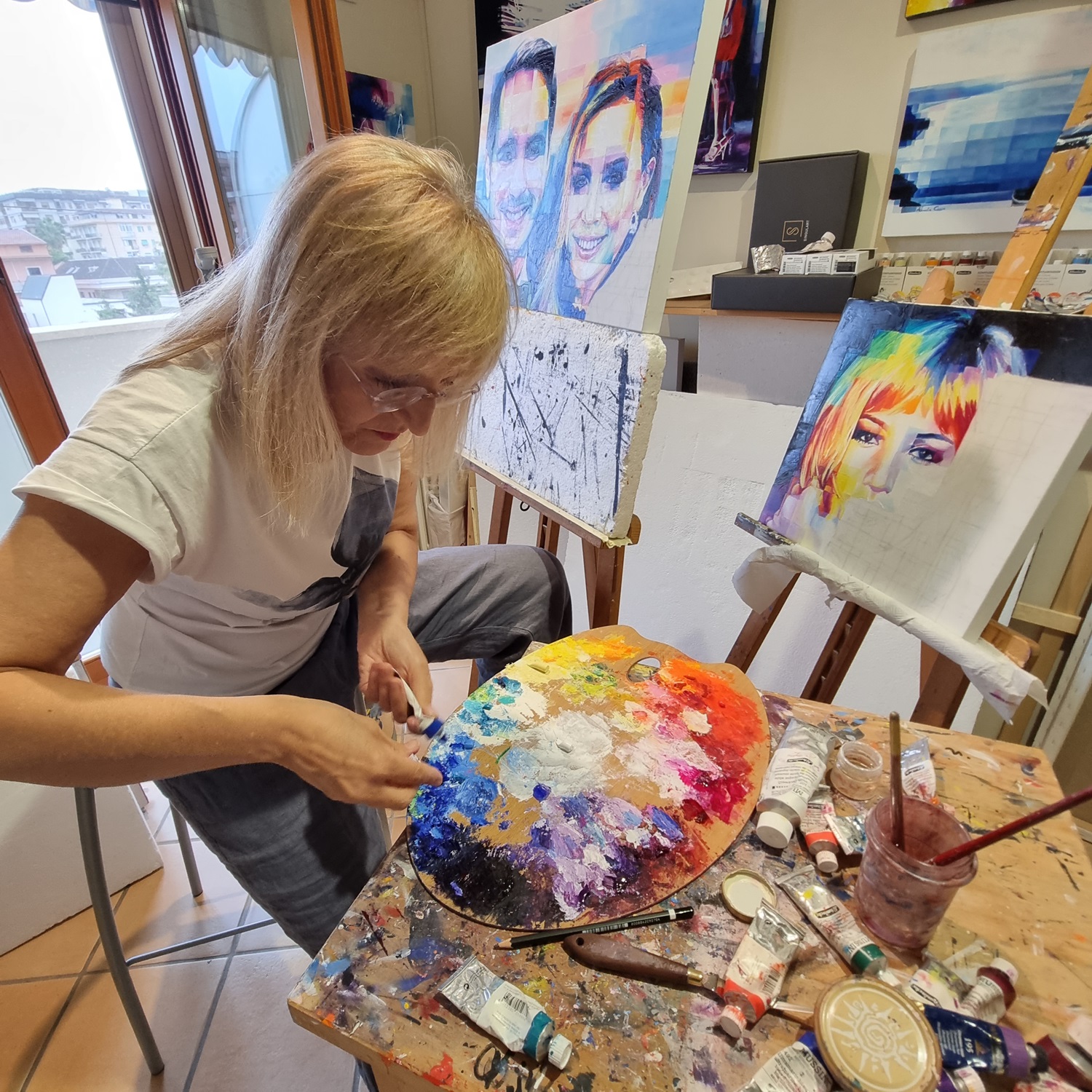 An artist working in a bright studio, blending oil paints on a colorful palette. Surrounding her are easels with in-progress portraits, featuring vibrant, pixelated designs of faces. The scene showcases the creative process, with tubes of paint, brushes, and tools scattered across the workspace, symbolizing the dedication and energy behind artistic expression.