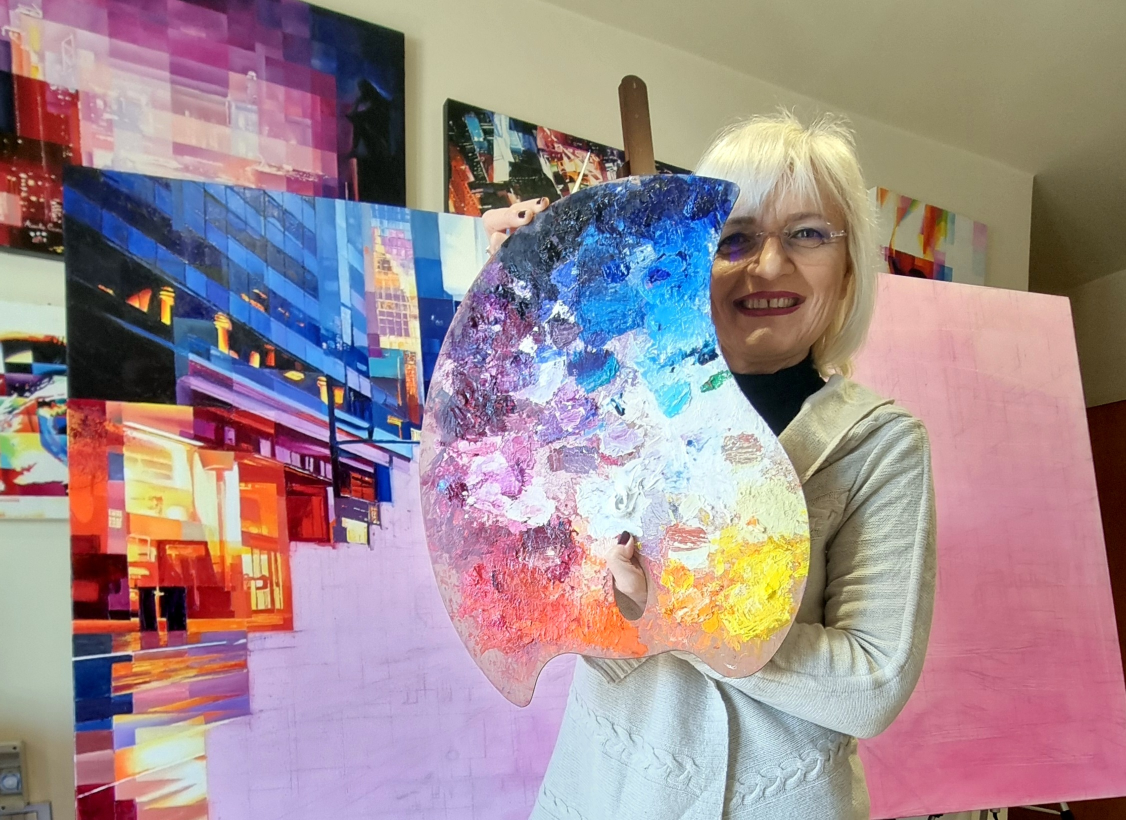 Artist Assunta Cassa in her studio, holding her colourful palette with Metropolitan series paintings in the background.